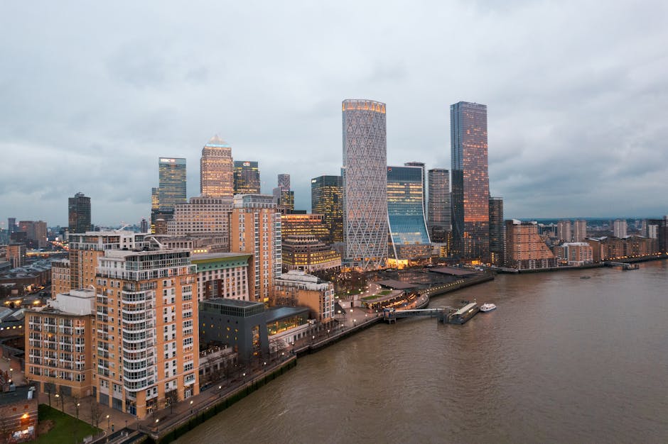 A panoramic view of Canary Wharf in London during dusk, featuring a cluster of modern high-rise office buildings with glass and steel facades reflecting the fading daylight. The scene includes a river