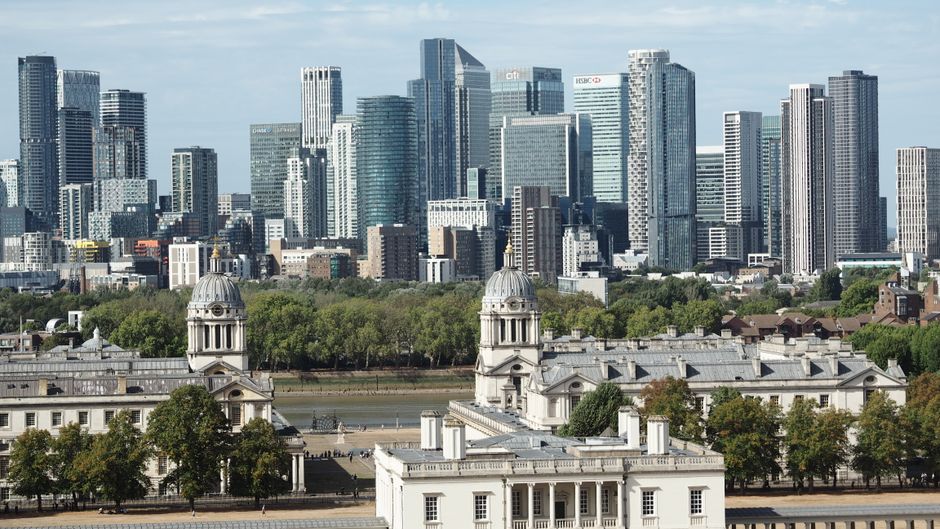 A panoramic view of Canary Wharf in London during dusk, featuring a cluster of modern high-rise office buildings with glass and steel facades reflecting the fading daylight. The scene includes a river