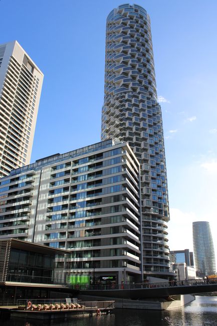 A tall residential building with a unique cylindrical design featuring multiple irregularly shaped balconies, situated among other high-rise structures under a clear blue sky. The building's exterior 