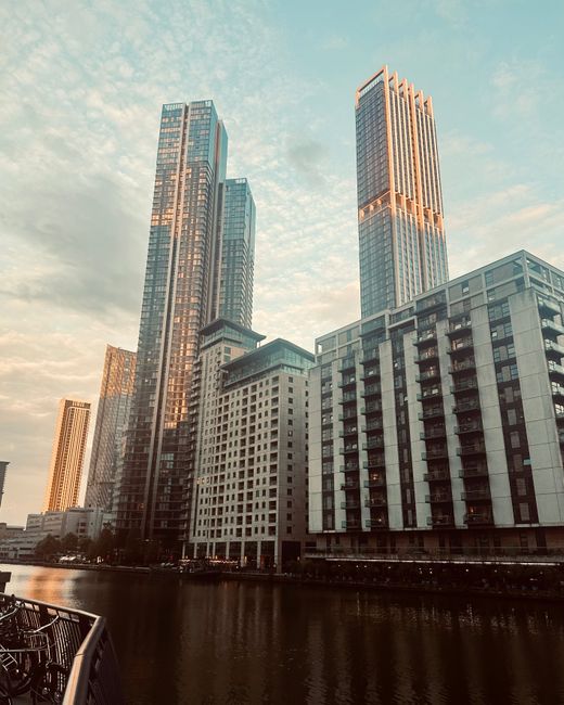 A black-and-white city skyline featuring a row of modern high-rise buildings constructed with glass and steel, situated along a waterfront. The buildings vary in height, with some showcasing sleek, re