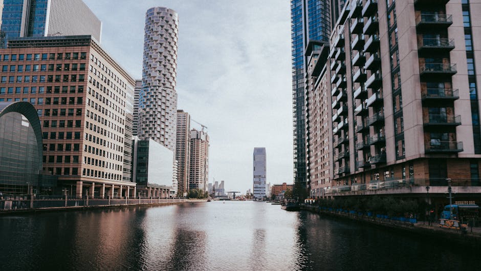 A panoramic view of Canary Wharf in the early evening, showcasing a cluster of modern high-rise office buildings with glass facades reflecting the sunset. The skyline includes several skyscrapers in v