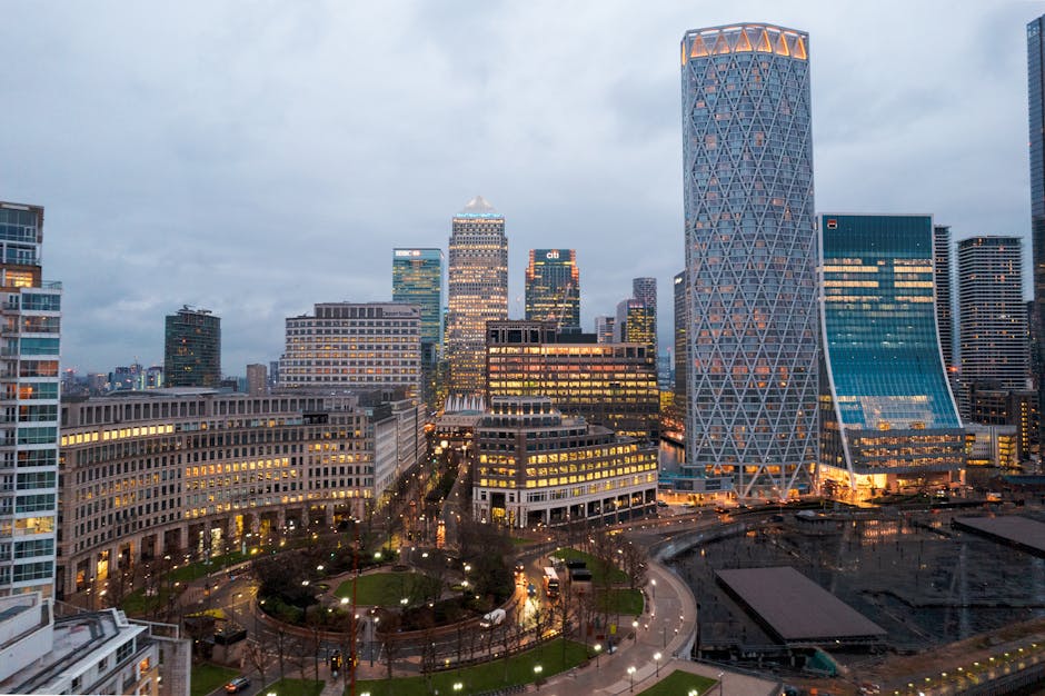A modern view of London's Canary Wharf skyline during daytime featuring a cityscape of tall glass and steel office buildings, including a prominent curved skyscraper and a building with a lattice faca